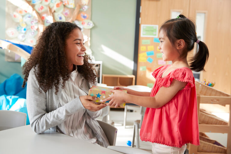 Kindergarten schoolgirl giving a gift to her female teacher in a classroom, side view, close up Kindergarten schoolgirl giving a gift to her female teacher